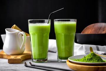 Matcha green tea powder latte with milk in two high glass on stone table, black straw, wooden plate and tray,bamboo tasaku and whisk