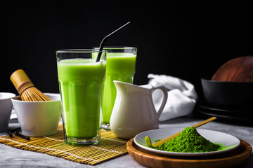 Matcha green tea powder latte with milk in two high glass on stone table, black straw, wooden plate...