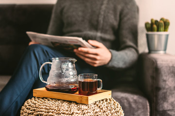 A young man in glasses, a gray sweater and blue trousers sits on a gray sofa ,reading a newspaper and drinking fresh hot coffee,Scandinavian-style Interior,Morning business Breakfast