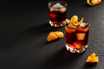 Negroni alcoholic cocktail in two glasses decorated with orange peel with ice cubes on a dark table