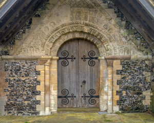 Old wooden door of a church.The wall has carvings over the archway.Part of the wall is made of Flint.