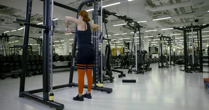 Sports girl in the gym doing standing exercises on the triceps, press with a barbell, raises and lowers the barbell, shakes his hands and shoulders. The view from the back.