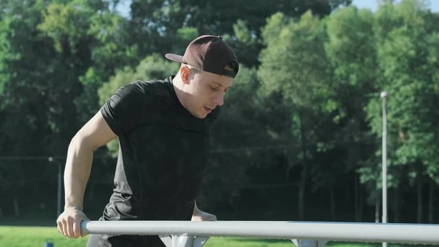 Young man in cap doing triceps dips on parallel bars at sunny morning. Side view of sporty male training in city park at summer. Man bars workout. 4k, UHD. Medium shot