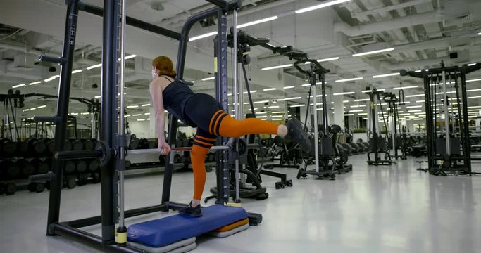 A sporty girl in the gym does a deadlift with a barbell and lunges and shakes her buttocks, a view from the back.