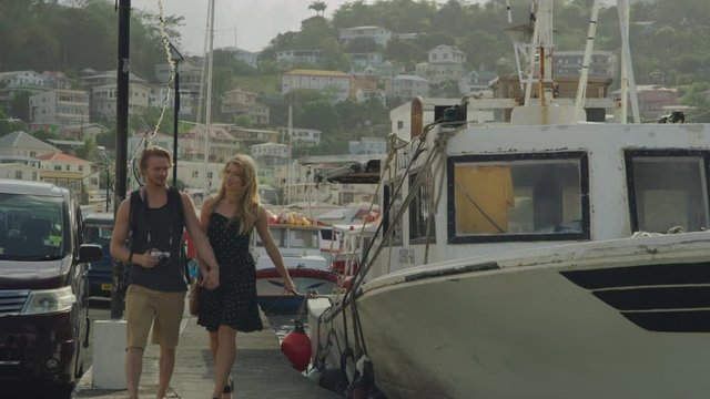 Approaching tourist couple holding hands and walking on waterfront near boat / St. Georges, Grenada