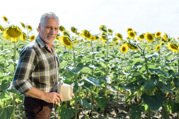 Fototapeta premium cheerful bearded farmer holding straw hat near field with sunflowers