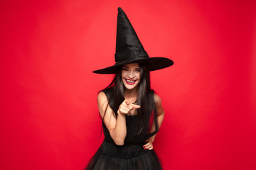 Young brunette woman in black hat and costume on red background. Attractive caucasian female model. Halloween, black friday, cyber monday, sales, autumn concept. Copyspace. Pointing, showing, choosing