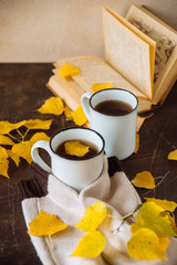 two white cups with tea in yellow leaves with a book and a knitted scarf, the concept of warmth and autumn sadness