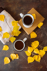 two white mugs surrounded by yellow leaves with a book and a knitted scarf, the concept of warmth and autumn sadness