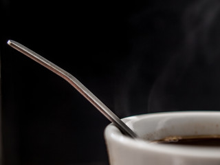 A piece of white cup with hot coffee, metallic spoon and smoke above it on the black background.