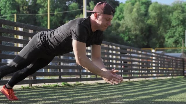 Young man doing push ups with clapping hands while exercise on green stadium. Medium shot of caucasian man doing push-ups outdoors in slow motion. Sport concept at sunrise. Workout