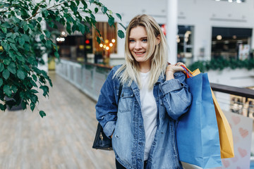 Time for shopping and spending time in trade center. Pretty woman in a denim jacket standing in a shopping mall with coloured shopping bags in hands. Shopping, sale, gifts concept, copy space