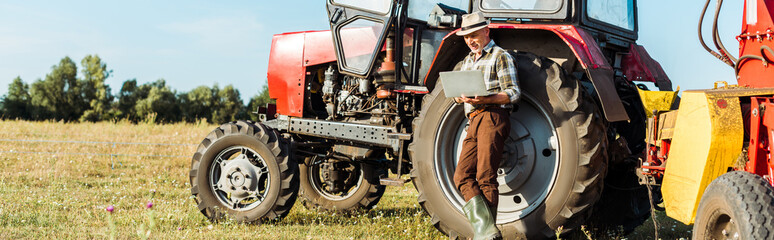 panoramic shot of farmer in straw hat using laptop near tractor
