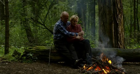 A love couple of two old people are in the forest and they are sitting near a burning fire on a log, hugging and talking.