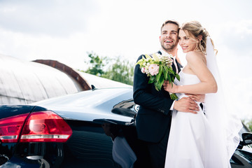 handsome bridegroom in suit hugging attractive and blonde bride with bouquet