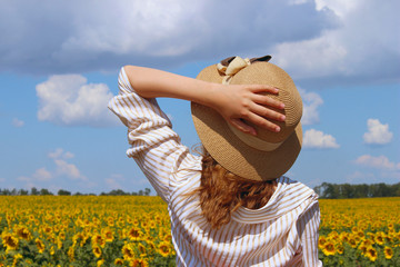 Cropped shot of a young girl, wearing a hat and white striped shirt, standing and looking to the yellow meadow over blue sky background. Young girl outdoors. People, Travel, Nature concept.