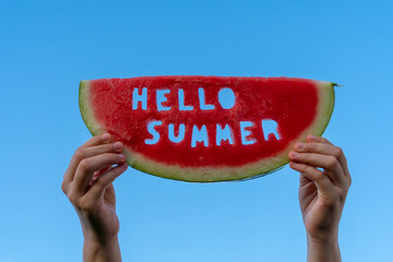 Fototapeta A piece of watermelon against a blue sky. Children's hands are holding a slice of watermelon with the text Hello Summer. Summer time concept obraz