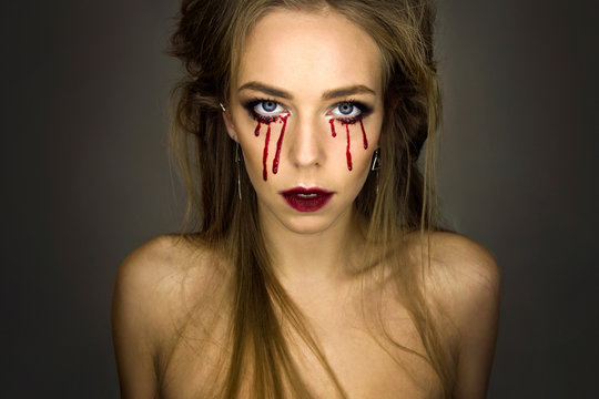 Close-up portrait of young stylish sexy blonde girl with dark bloody makeup with blood drops from eyes which is standing and looking straight on gray background, halloween concept, free space
