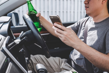 Young asian man drives a car with drunk a bottle of beer and using smartphone behind the wheel of a car