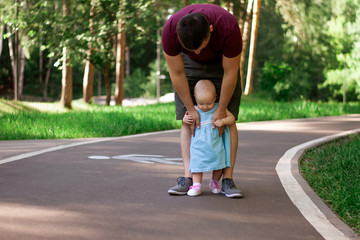 Dad teaches daughter to walk. Happy dad and daughter. Dad with a one-year-old daughter in a dress are walking in the park. Teaching children, fatherhood. The first steps.