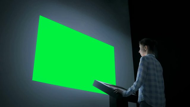 Woman looking at blank large interactive wall green display in dark room of modern technology exhibition or museum. Mock up, green screen, presentation, template, education, chroma key concept