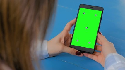 Woman looking at black smartphone mobile device with blank green display on blue table at home - over shoulder close up view. Mock up, chroma key, copyspace, green screen, template, technology concept