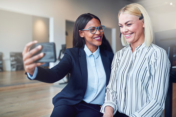 Two young businesswomen laughing while taking selfies in an offi