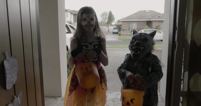 a front door opens to two young children, a boy and a girl, carrying trick or treat buckets and wearing werewolf and day of the dead costumes for Halloween 