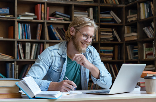 Smiling male student using laptop looking at computer screen learn easy internet course study online e learning in app write notes prepare for test exam watching educational video sit at library desk