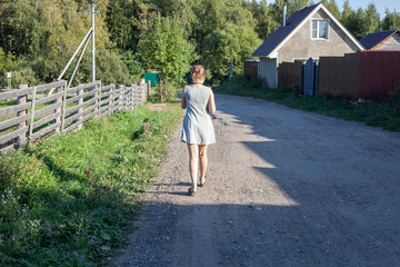 A girl walks along the fence of the farm. A girl walks out of town on a highway. Girl in the summer in a light dress.