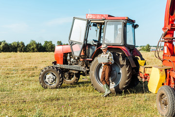 bearded farmer in straw hat using laptop near tractor