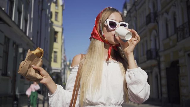Positive beautiful young woman walking on the street drinking coffee and eating croissant. Attractive fashionable girl enjoying sunny day in the old European city. Tourism concept