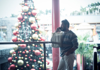 Cheerful adult caucasian man phone calilng in commercial center with christmas tree and lights in background - xmas time in the city concept for happy people