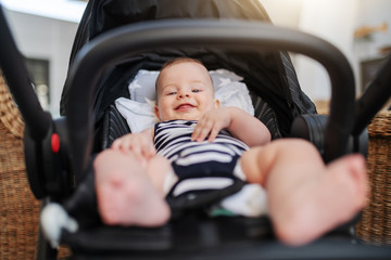 Low angle view of playful Caucasian smiling baby boy 6 months old dressed in striped baby clothes sitting in stroller and looking up.