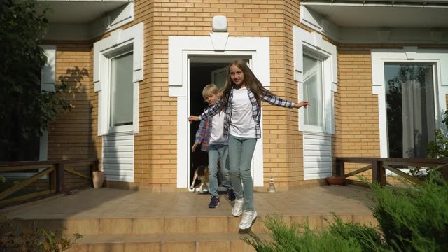 Little cute boy and girl with backpacks leaving the house and going to school. Smiling mother waving hand. Happy loving family concept