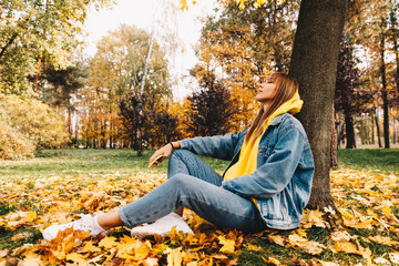 Autumn walk. Technologies. Happy girl in jean clothes is holding a smartphone and enjoying the weather with her eyes closed while sitting on the ground in the park