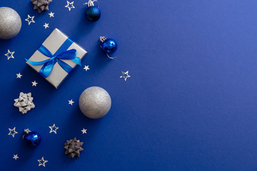 Christmas silver gift box with bow ribbon, baubles, decorations, confetti on magic dark blue background with copy space. Vintage Xmas greeting card mockup, postcard template. Flat lay, top view.