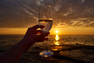 hand holding glass of white wine and with sea and beautiful sunset at background, close-up. Summer evening relaxed mood concept