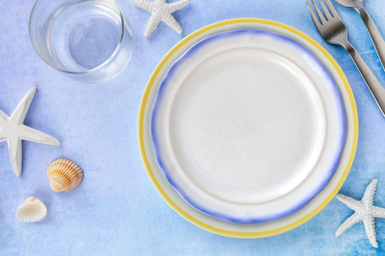 Table setting for kids with an empty plate, a fork and a transparent glass on a light blue table with marine themed decorations as a summer or holiday concept