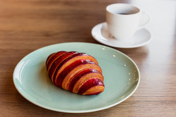 A horizontal photo of a croissant with strawberry filling on a bright green plate and a white cup of black coffee.