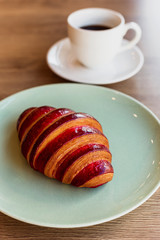 A vertical photo of a croissant with strawberry filling on a bright green plate and a white cup of black coffee.