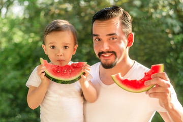 Father and son together eating watermelons, both man and kid are holding slices of juicy watermelons, drops of juice on face of kid. Green leaves in background. Family meal outdoors in sunny evening