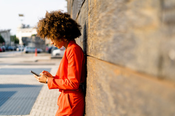 Young woman wearing fashionable red pantsuit leaning against wall using cell phone