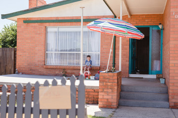 Boy sitting on front porch of home with toy sale sign on fence