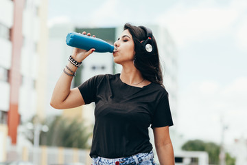 Beautiful young woman using headphone listening to music and drinking water outdoors