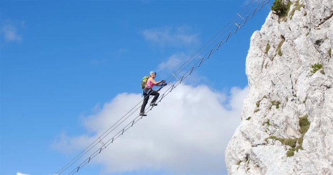 Woman moving high on a via ferrata diagonal ladder on Donnerkogel mountain near Gosau, against the blue sky and white clouds. Stairway to heaven concept.