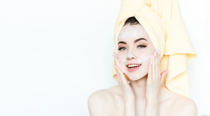 Fototapeta premium Portrait of cheerful woman applying foam for washing on her face. Beautiful model with towel on head with healthy glow perfect smooth skin. Young girl enjoys facial skincare routine.