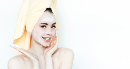 Naklejka premium Portrait of cheerful woman applying foam for washing on her face. Beautiful model with towel on head with healthy glow perfect smooth skin. Young girl enjoys facial skincare routine.