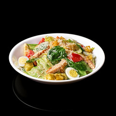 Tasty appetizing fresh salad Caesar with chicken, tomatoes, the eggand cheese parmesan in bowl on black background. View from above.