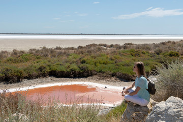 Young blond woman sitting on the rock next to colorful salt lagoon in Camargue Nature Park, Southern France - obrazy, fototapety, plakaty